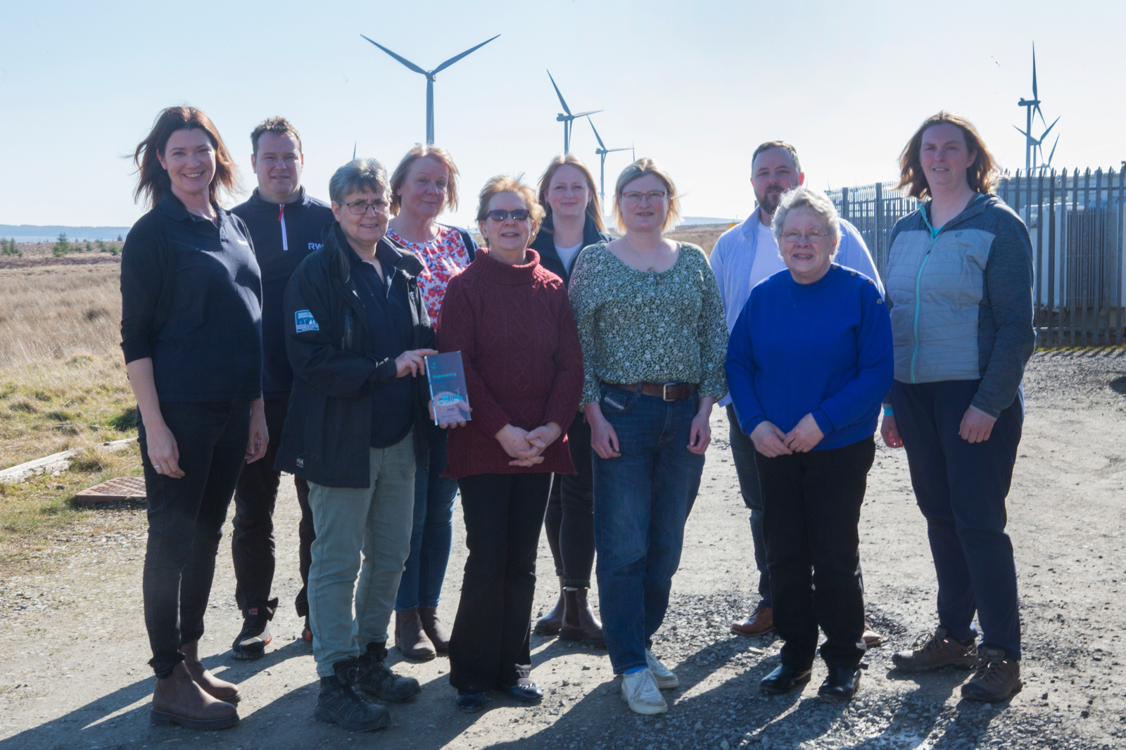 A group of individuals stands on a gravel path surrounded by wind turbines, smiling and engaged during a sunny day.