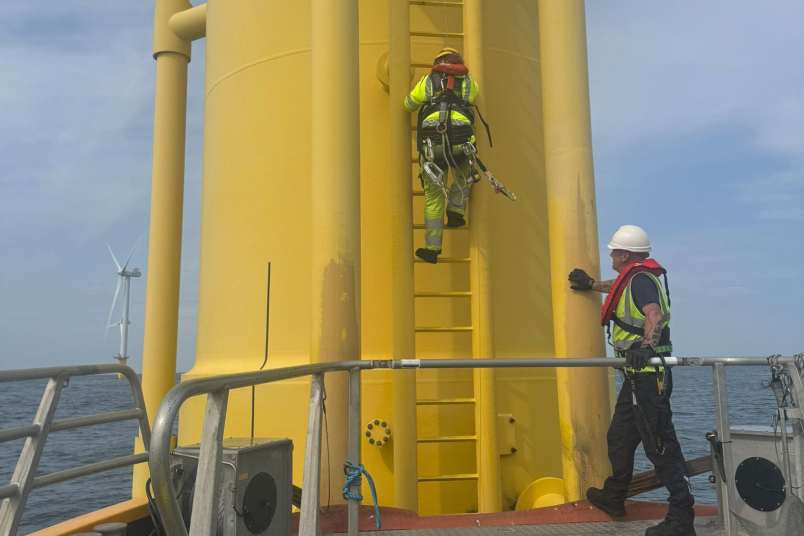 Two workers in safety gear are near a yellow structure at sea. One is climbing a ladder, while the other observes from below.
