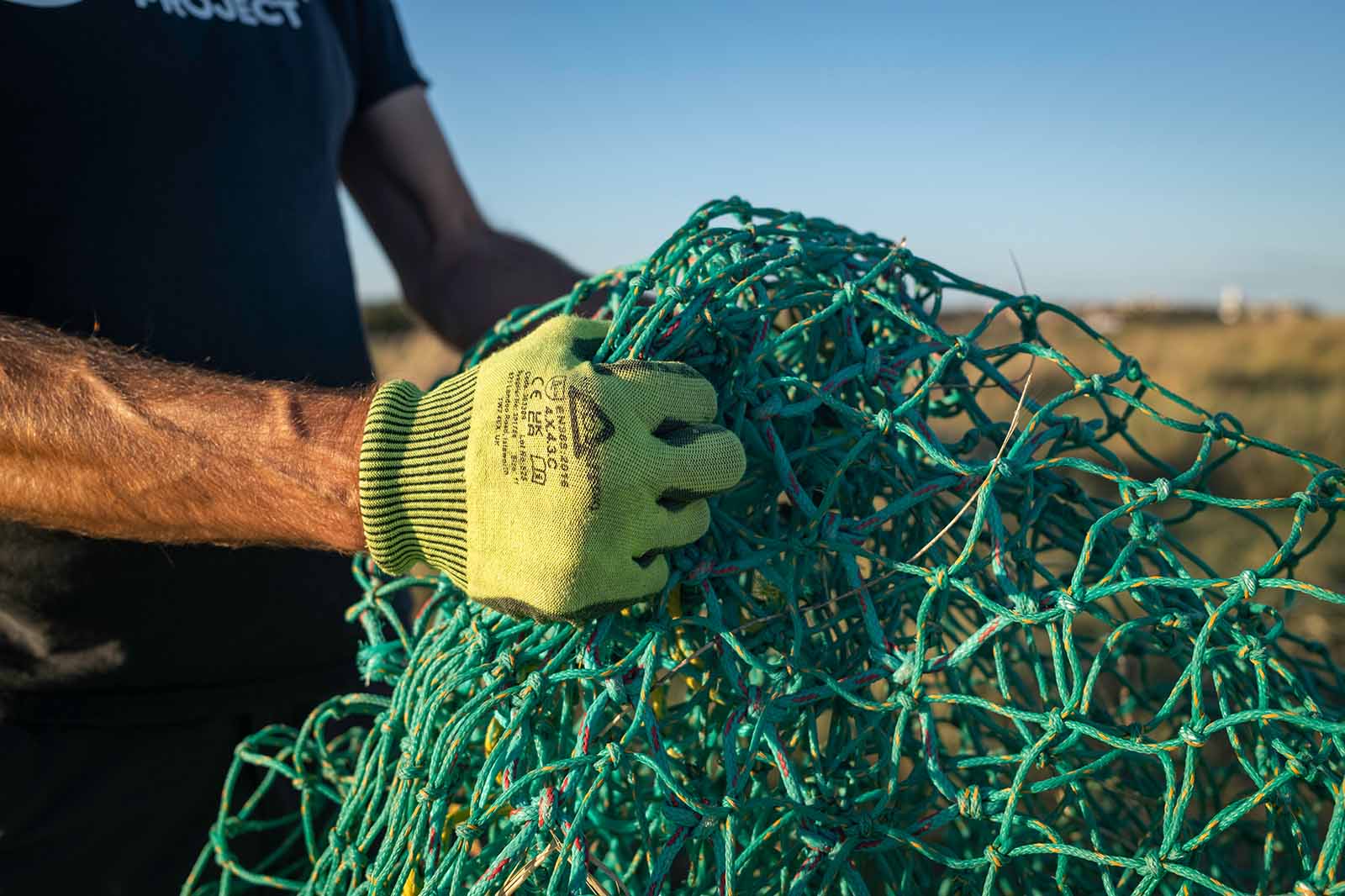 A person wearing a high-visibility green glove holds a tangled turquoise fishing net, with a tanned forearm visible in sunlight.