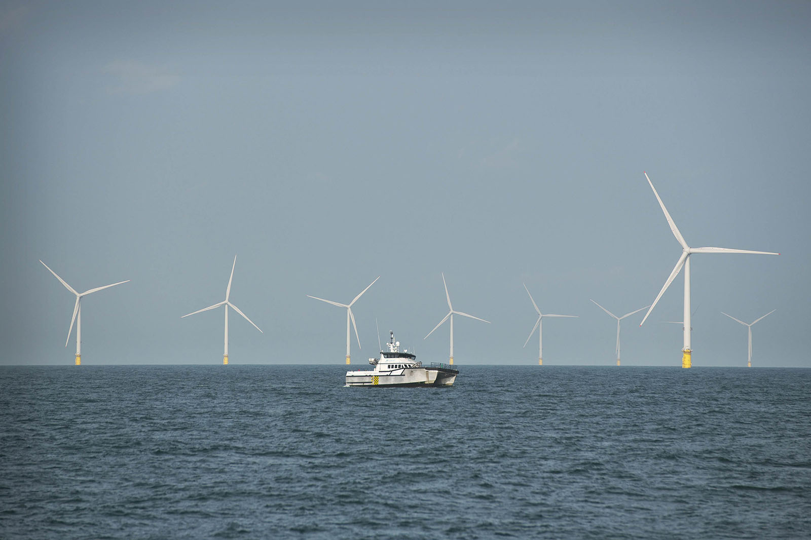 A crew boat passes in front of a row of large offshore wind turbines with yellow bases under a pale blue sky.