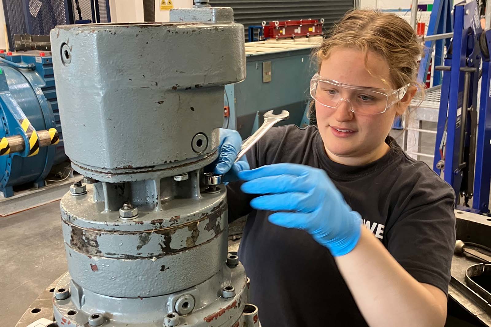 A person wearing blue nitrile gloves and a black shirt uses a wrench to tighten bolts on a large gray industrial gearbox in a workshop. Picture of Madeleine Warburton.