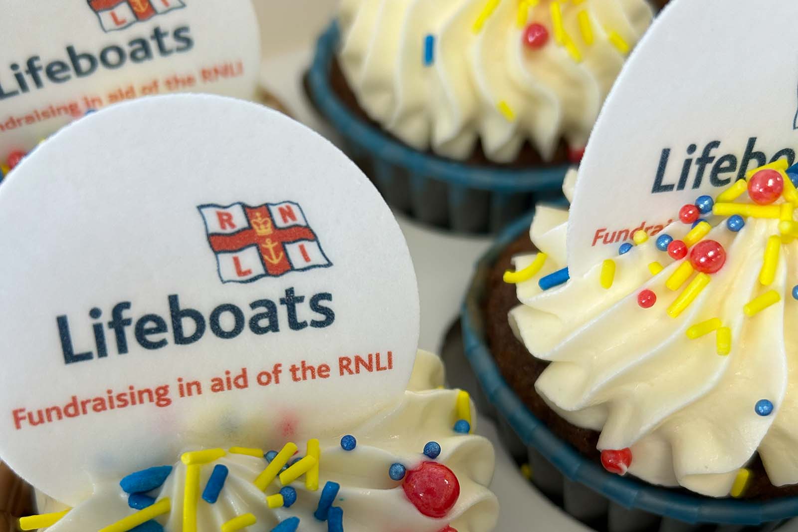 Chocolate cupcakes with swirled white buttercream, blue paper liners, yellow, blue and red sprinkles, and discs reading "Lifeboats" and "Fundraising in aid of the RNLI".