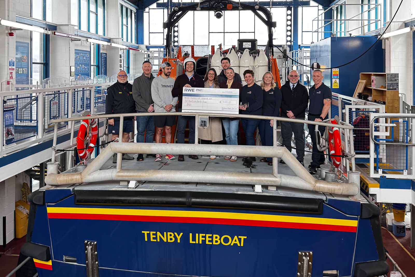 A smiling group stands on a raised platform inside a boathouse behind a dark blue lifeboat labeled "TENBY LIFEBOAT", holding a large cheque.