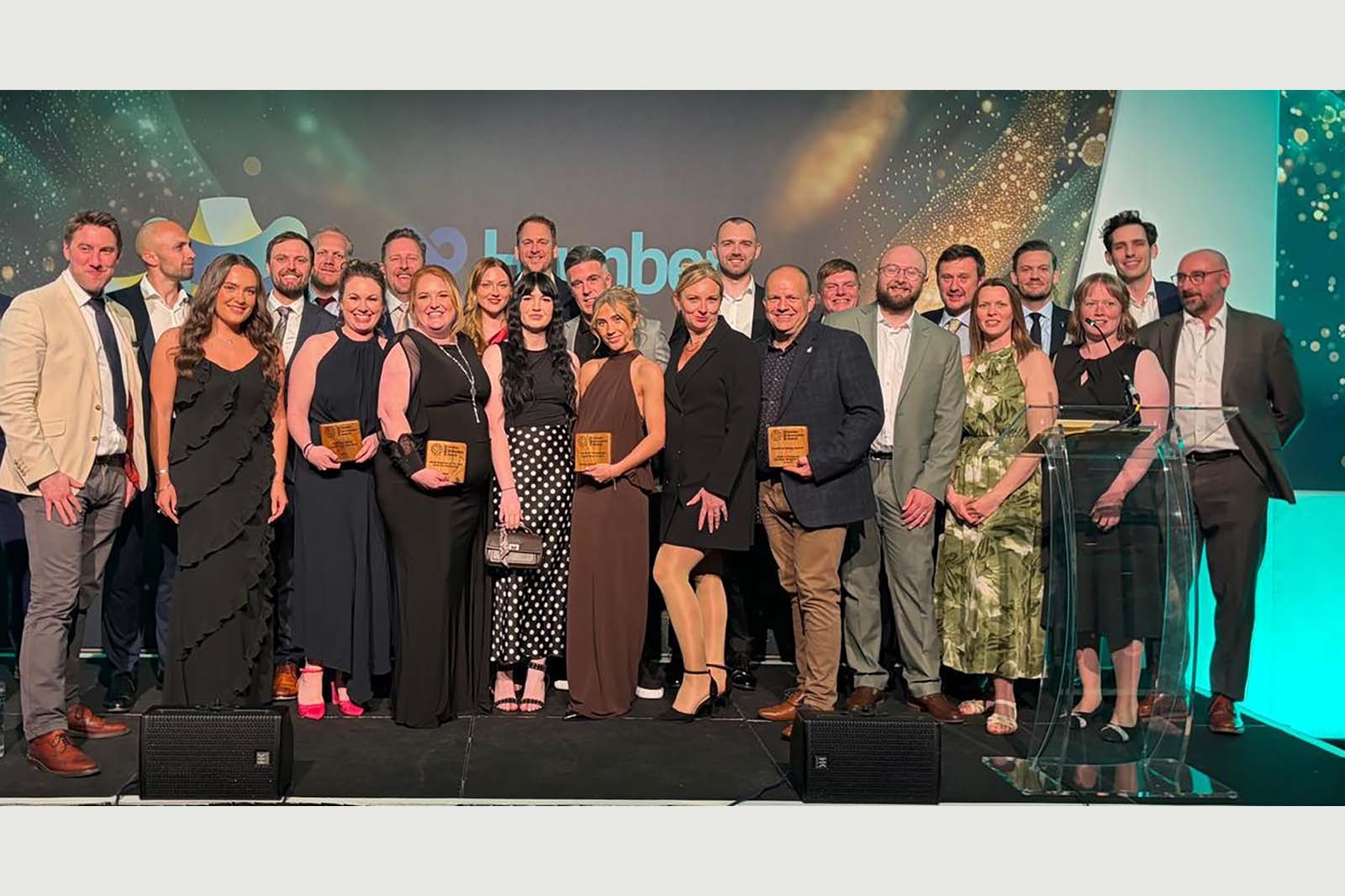 A group of about twenty formally dressed adults stand on a stage holding wooden plaque awards in front of a teal and gold glittering backdrop.