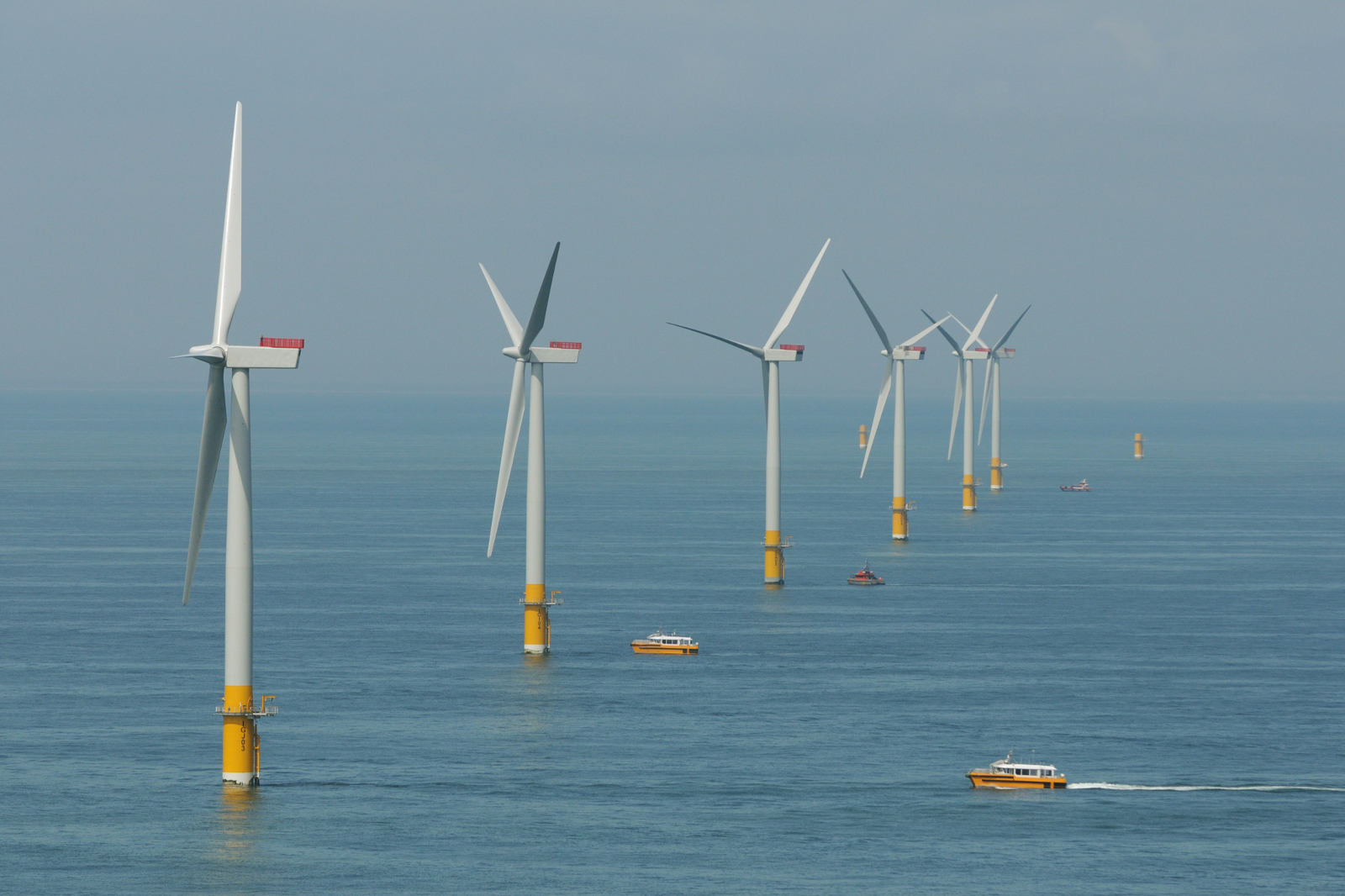 A line of white offshore wind turbines with yellow bases stands in calm blue sea beneath a pale sky, as orange service boats pass.