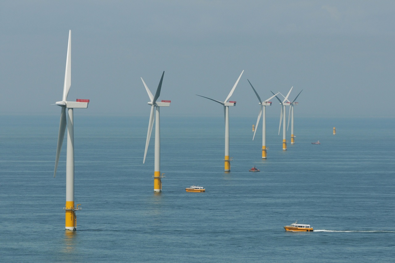 A line of white offshore wind turbines with yellow bases stands in calm blue sea beneath a pale sky, as orange service boats pass.
