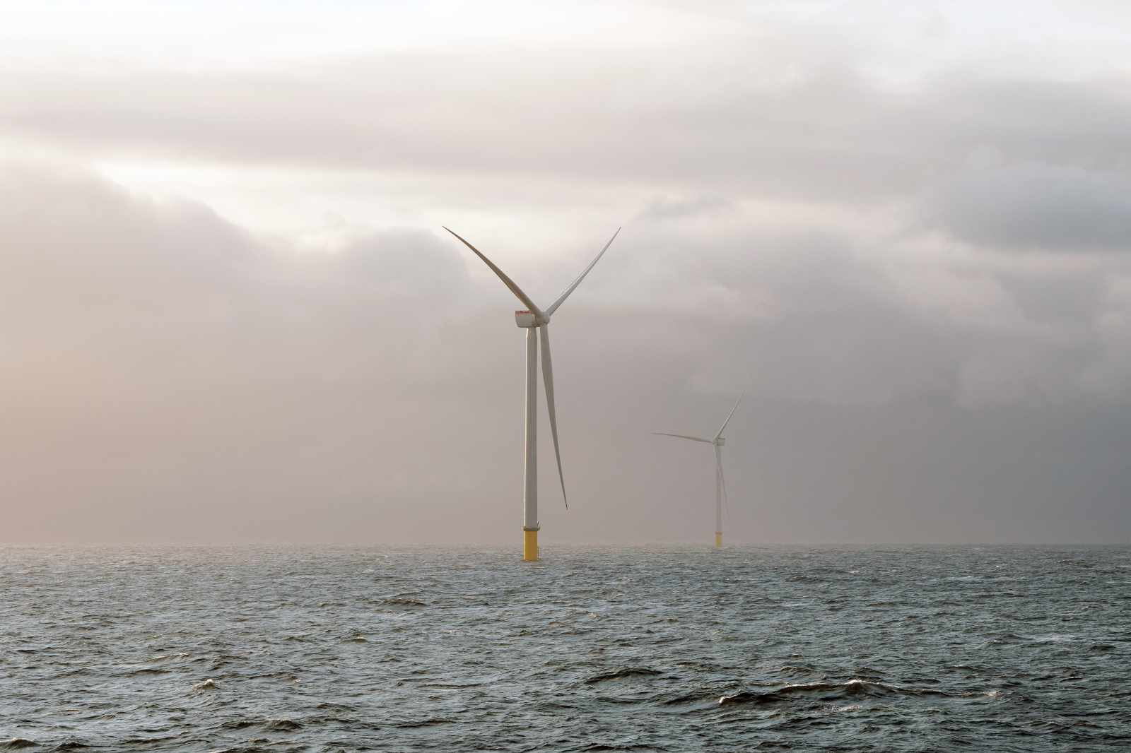 Two white offshore wind turbines with yellow bases rise from a grey sea beneath a soft, overcast sky with pale pink light.
