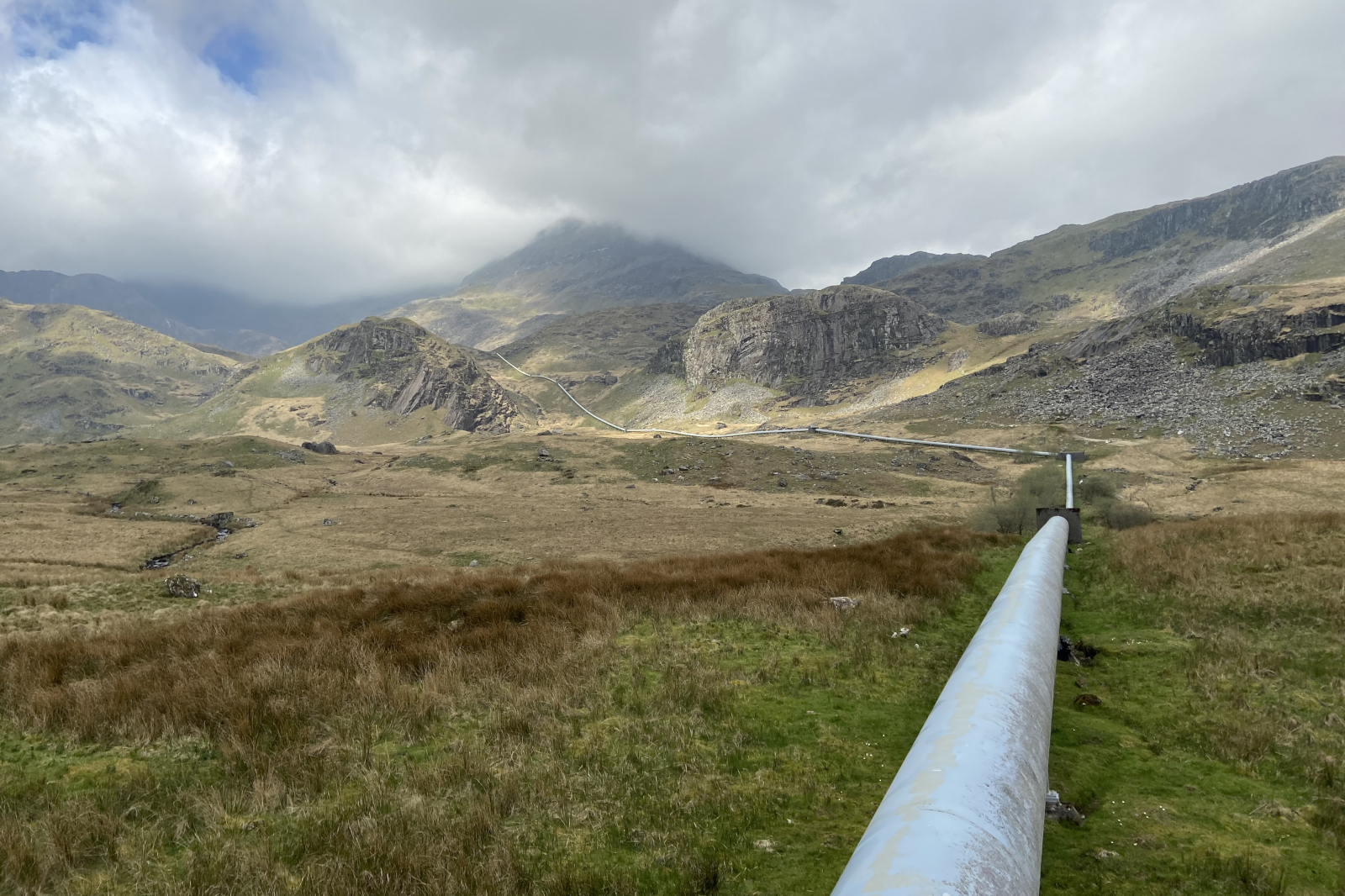 A pale blue metal pipeline runs diagonally across green-brown moorland toward rocky gray hills under low, brooding clouds.