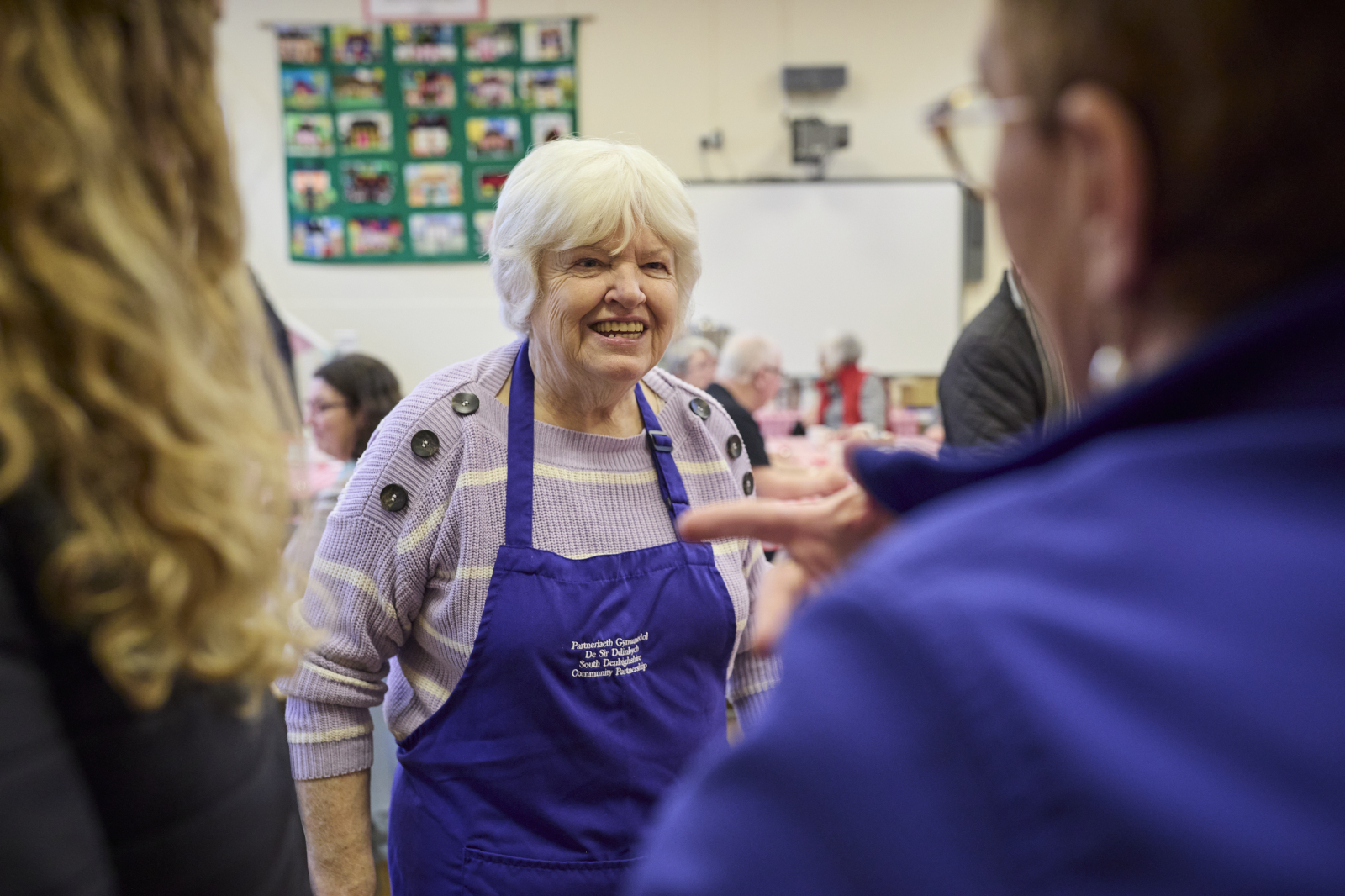 An elderly woman with white hair wears a purple apron over a striped sweater in a busy community room with a quilt on the wall.