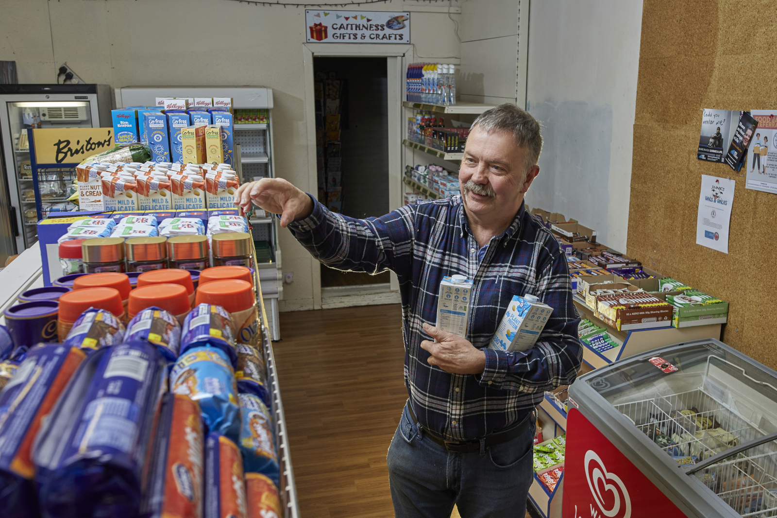 A person in a blue plaid shirt reaches for packaged goods on a white shelf while holding two cartons in a small shop.