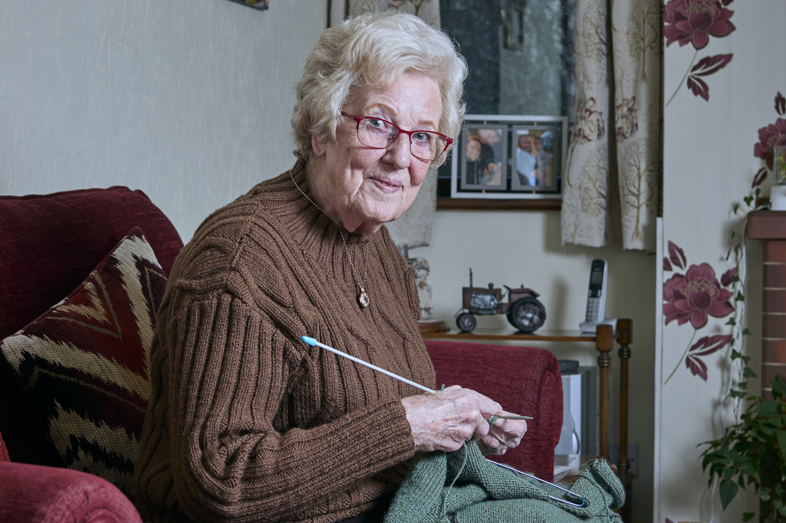 An elderly person with white hair sits knitting green yarn on a red armchair, wearing a brown sweater in a cozy living room.