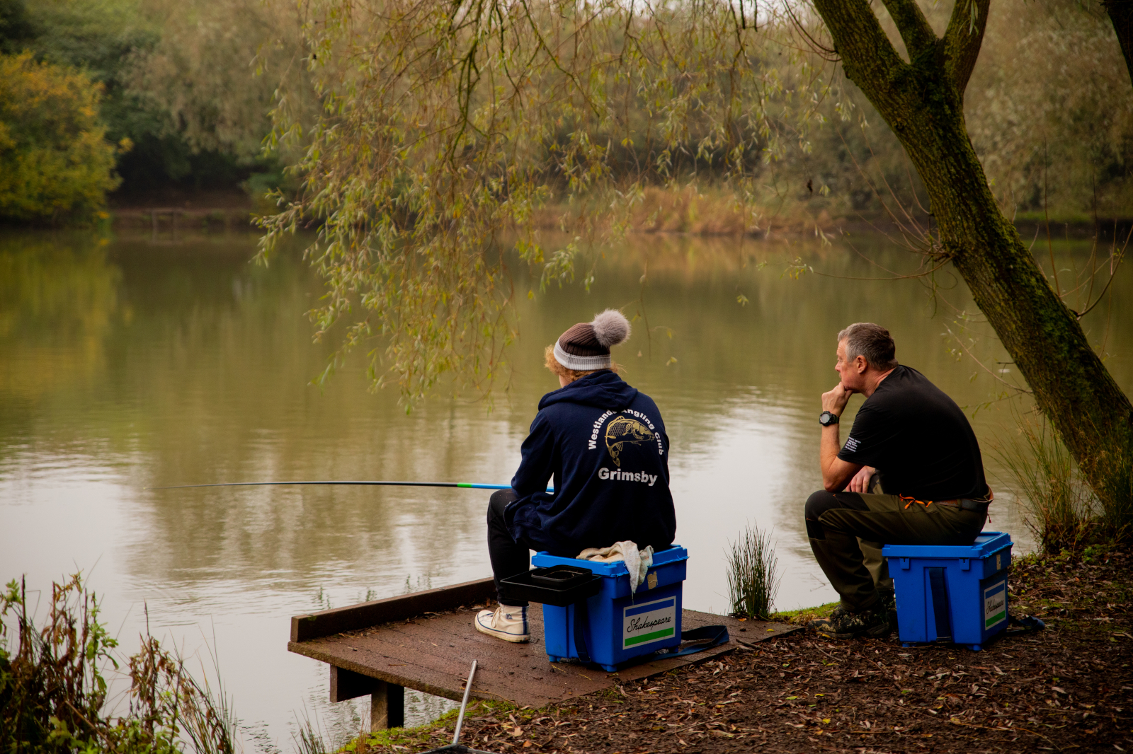 Two men sit on blue plastic boxes on a small wooden platform by a calm green lake under a leaning tree, one jacket labeled "Grimsby".