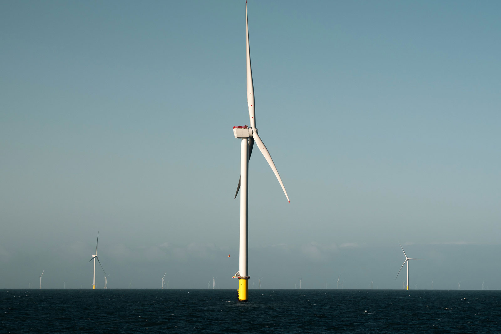 A large white offshore wind turbine with a yellow cylindrical base rises from a dark blue sea under a pale blue sky.