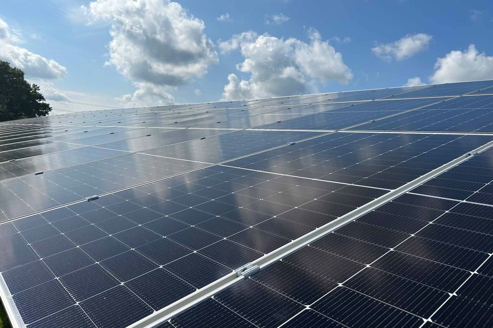 A wide array of dark photovoltaic solar panels with aluminum mounting rails, their glass surfaces reflecting white clouds against a bright blue sky.