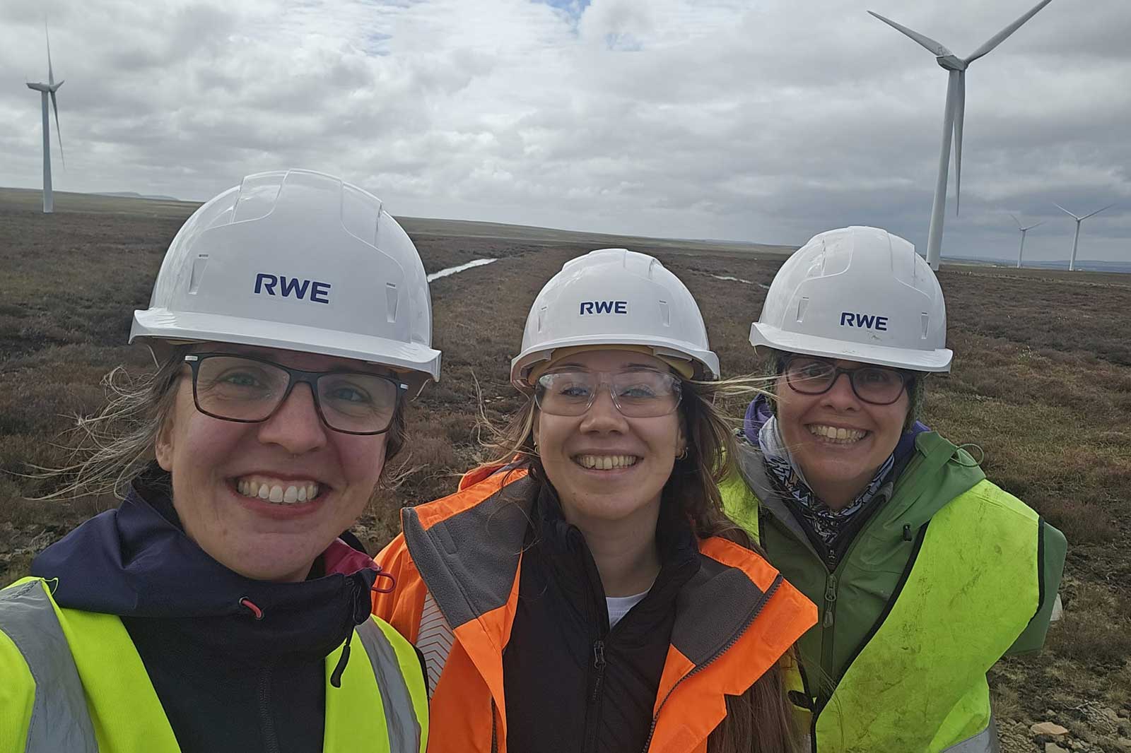 Three smiling people in white helmets labeled "RWE" and orange and yellow high-visibility vests stand on moorland with wind turbines under a cloudy sky.