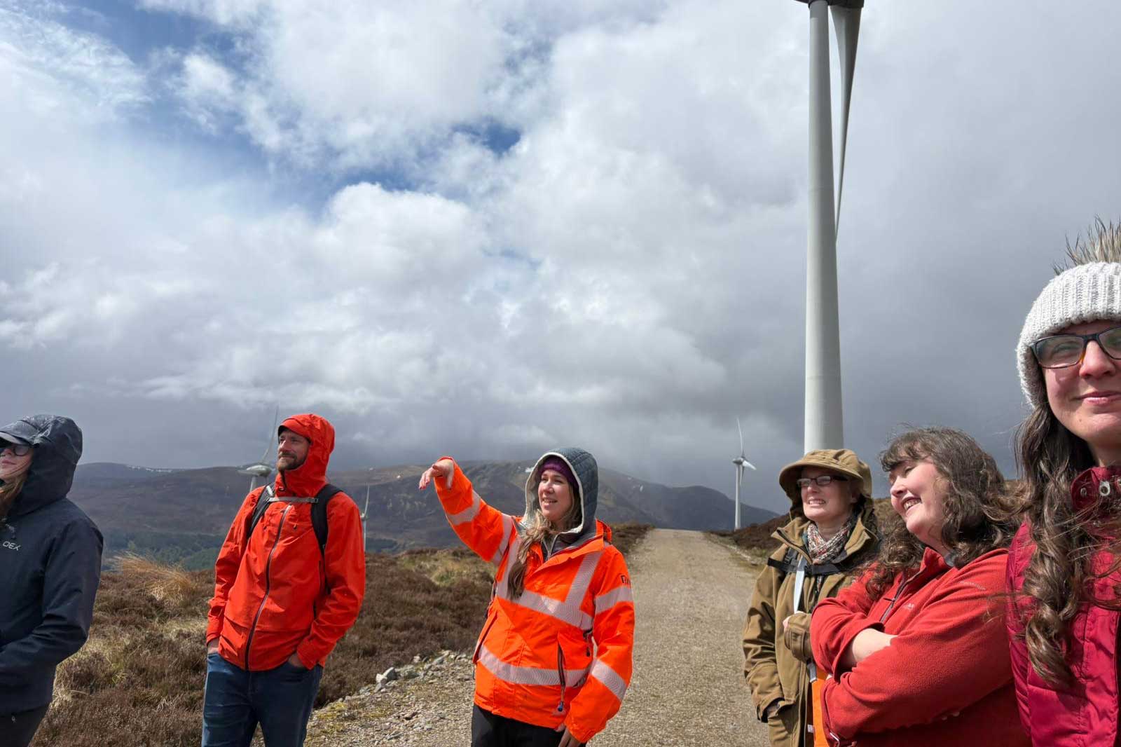 Six people in bright orange, red and khaki jackets stand on a gravel path beside a white wind turbine under cloudy skies.