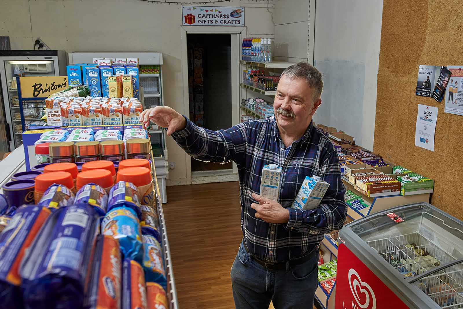 A smiling shopkeeper in a blue plaid shirt reaches for a packaged product while holding milk cartons beneath a sign reading "CAITHNESS GIFTS and CRAFTS".