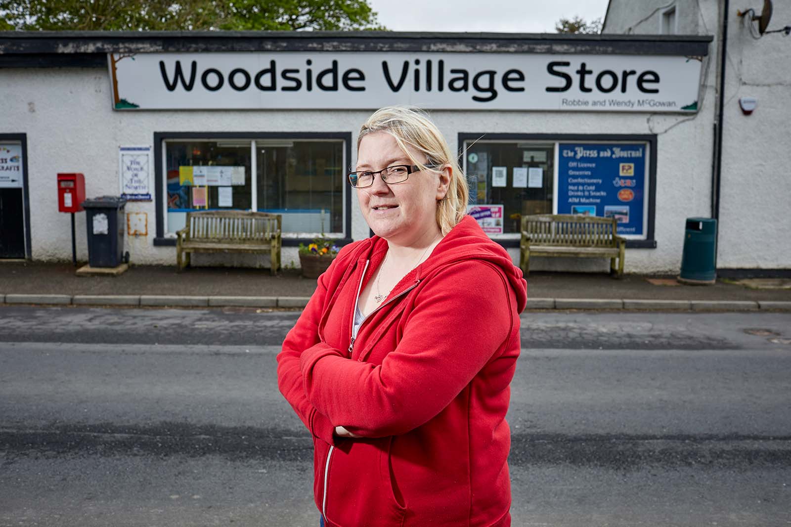 A woman in a red hoodie and glasses stands with arms crossed, smiling before the white shopfront labeled "Woodside Village Store".