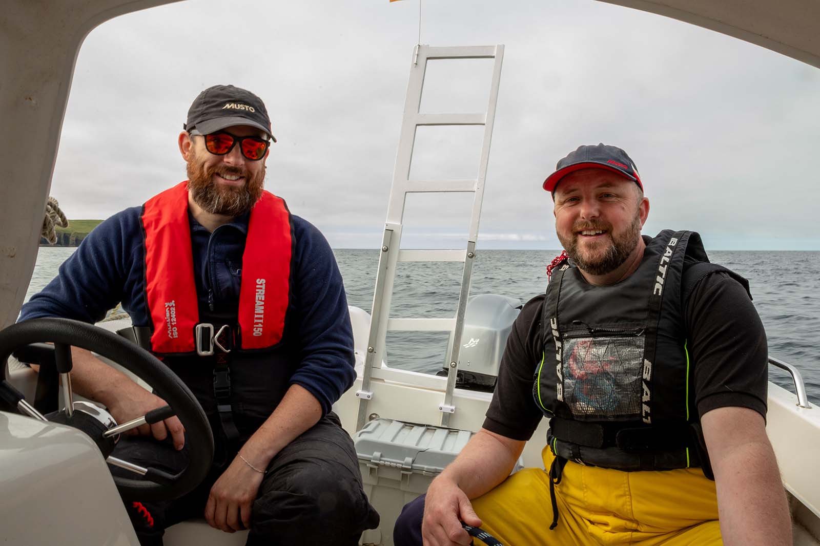 Two smiling bearded men sit in a small white motorboat wearing red and black life jackets, one steering and gray sea behind them.