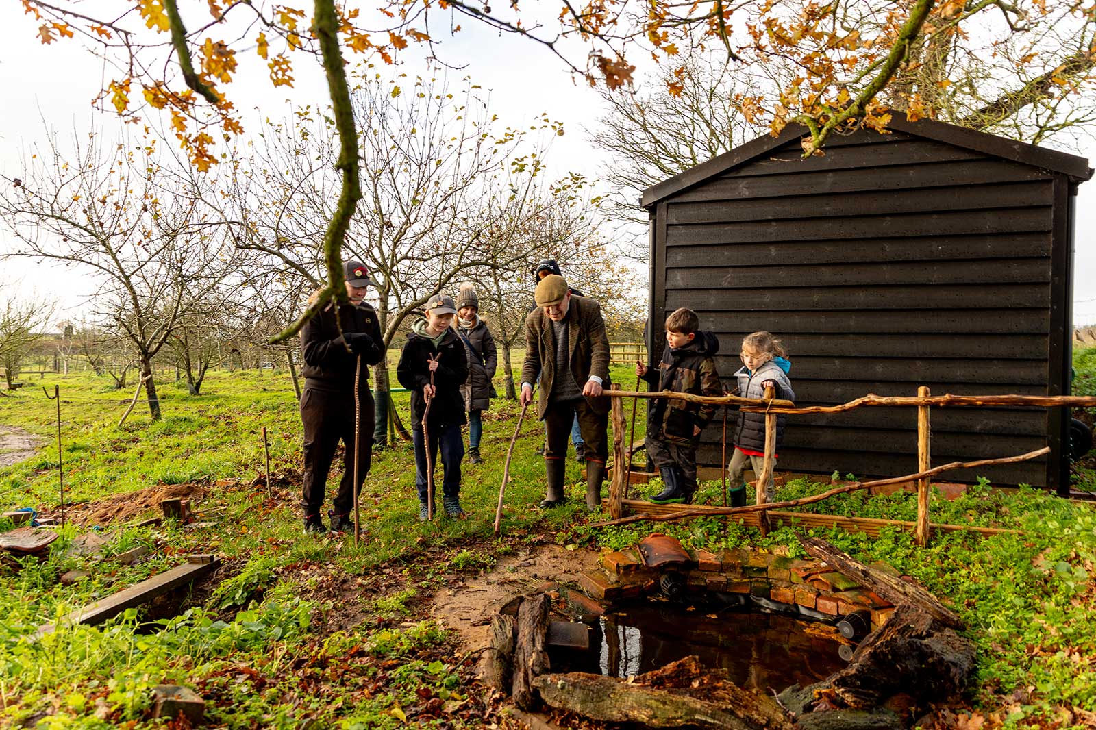 Six people in coats stand by a dark wooden shed and rustic fence beside a brick-lined puddle in a grassy autumn orchard.