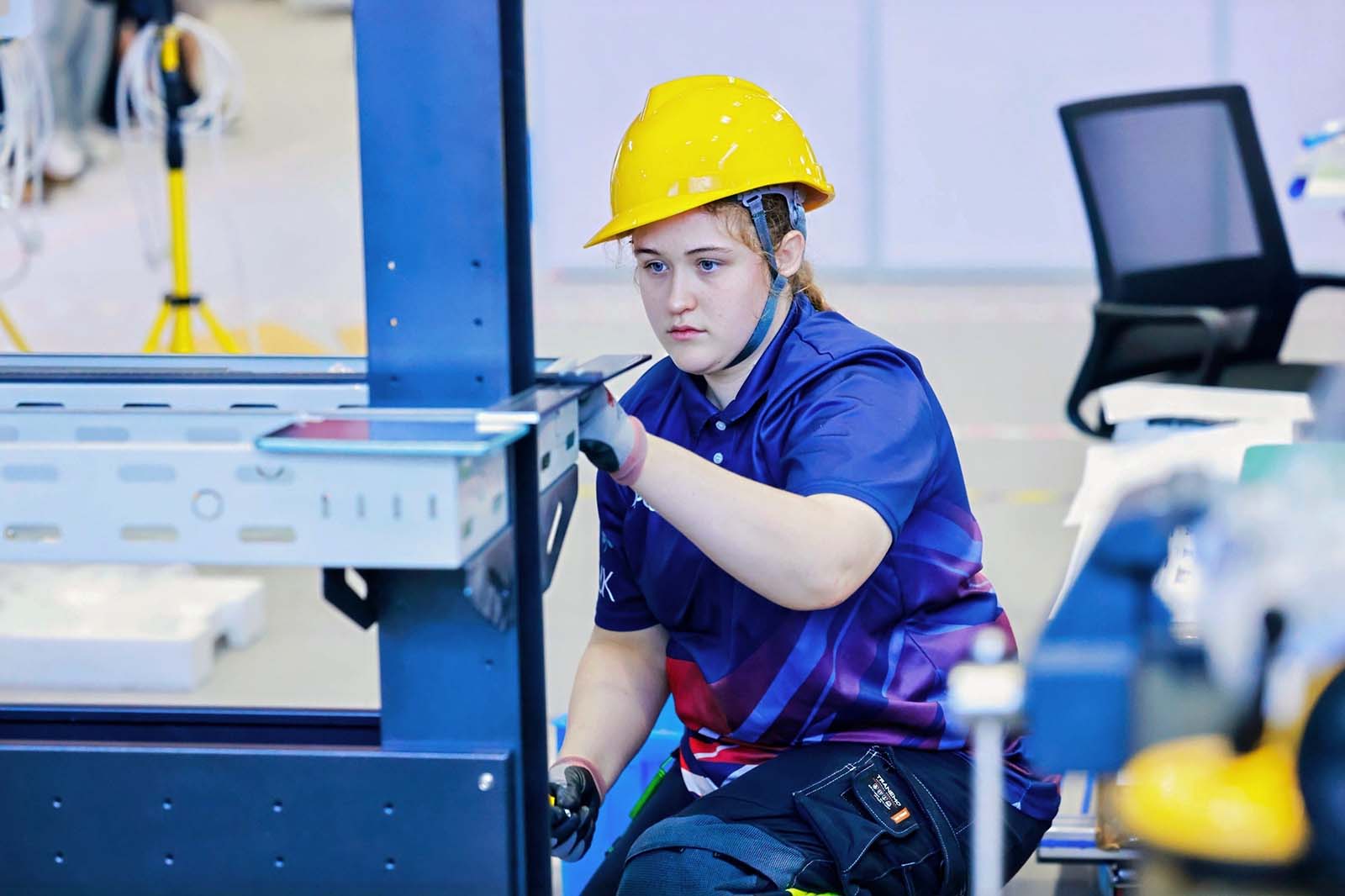 A young worker in a yellow hard hat and blue-and-purple polo kneels while assembling a dark metal frame in a bright workshop.