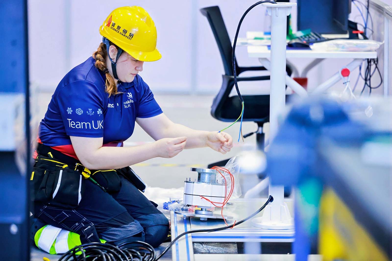 A woman kneeling at a workbench connects colored wires to a metal motor, wearing a yellow hard hat and a blue shirt labeled "Team UK".