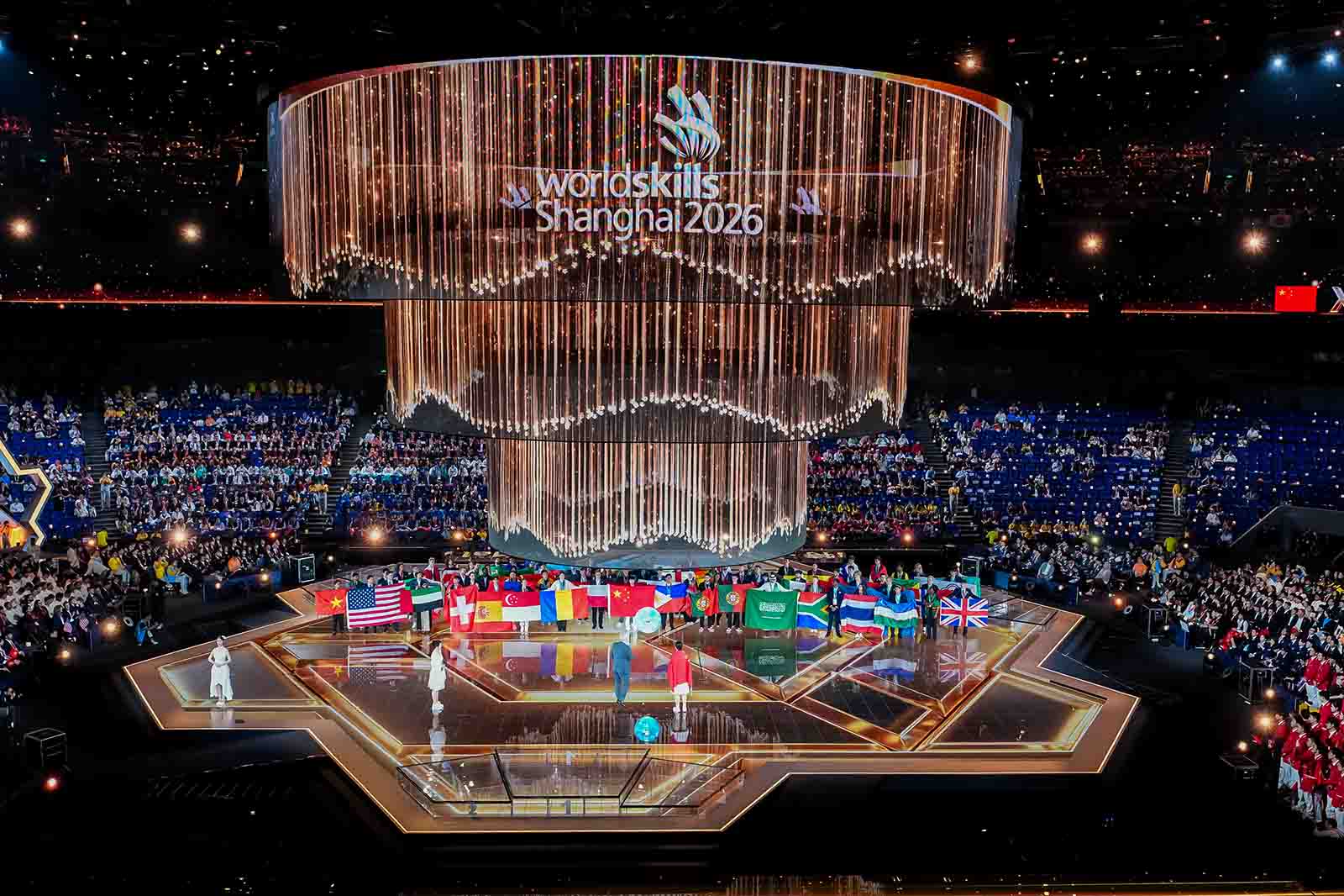 A star-shaped stage with flags and performers beneath a cascading cylindrical curtain of golden lights and the text "WorldSkills Shanghai2026" overhead, creating a festive atmosphere.