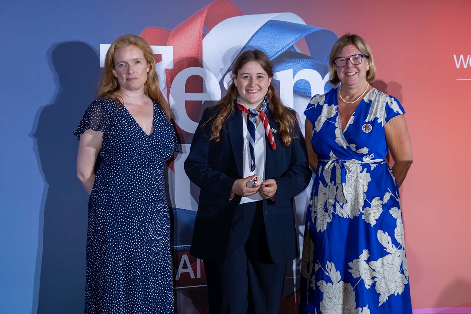 Young girl in a navy suit and scarf smiles between two women in blue dresses before a red, white and blue backdrop reading "Team".