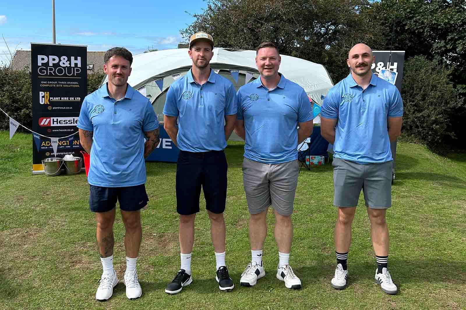 Four men stand on grass wearing matching light blue polo shirts and shorts, with a white canopy and a sign saying "PP&H GROUP" behind them.