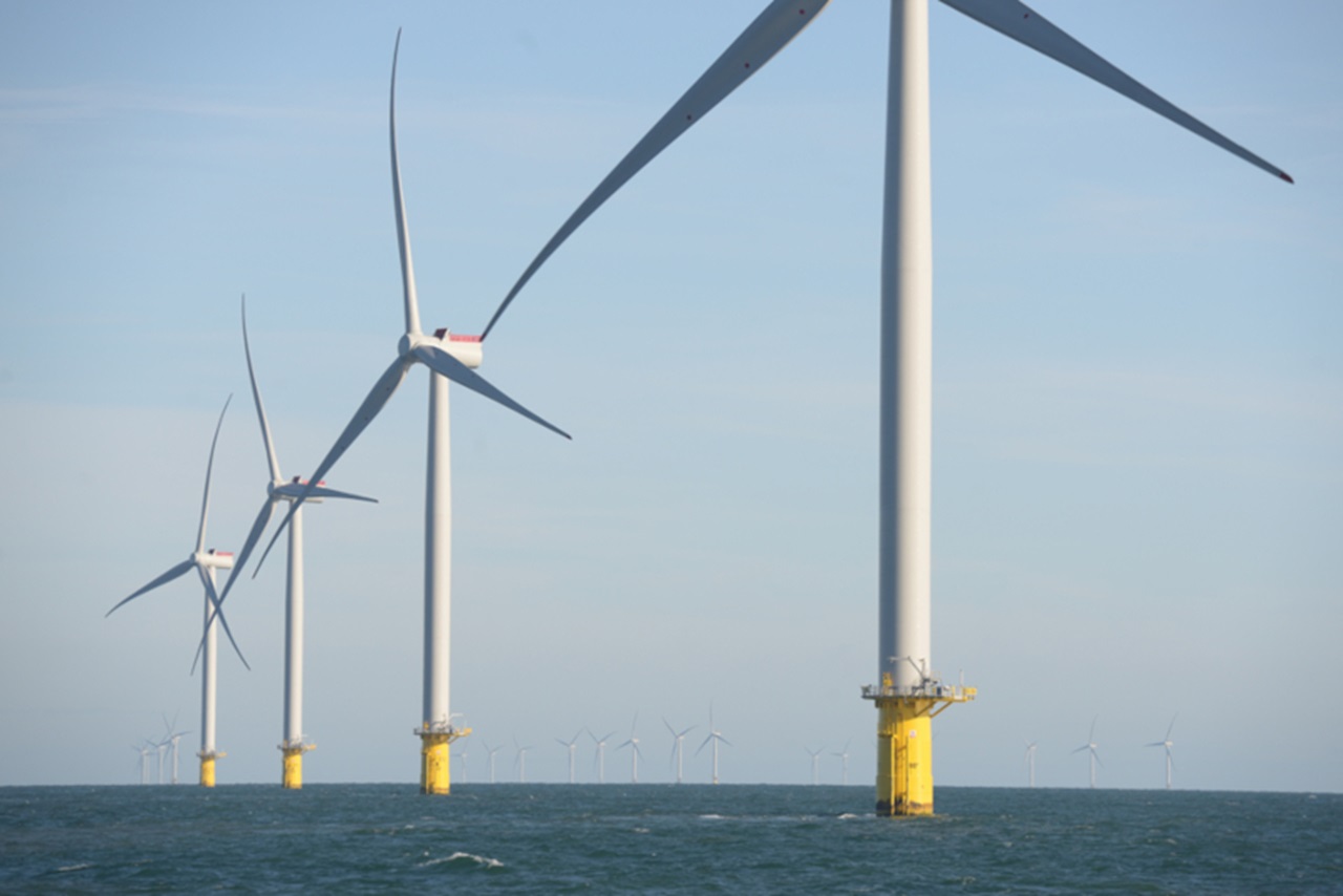 A line of white offshore wind turbines with yellow cylindrical bases rises from the calm blue sea under a pale sky.