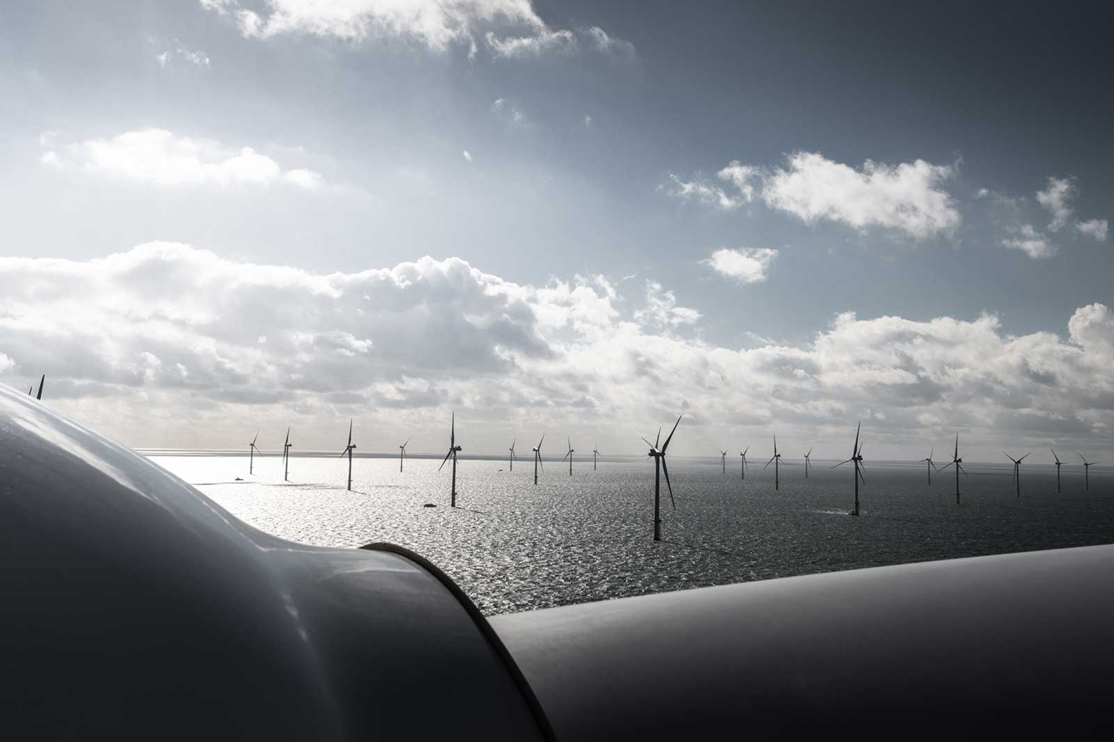 A view of offshore wind turbines in the distance, with a cloudy sky and sun reflecting on the water's surface.
