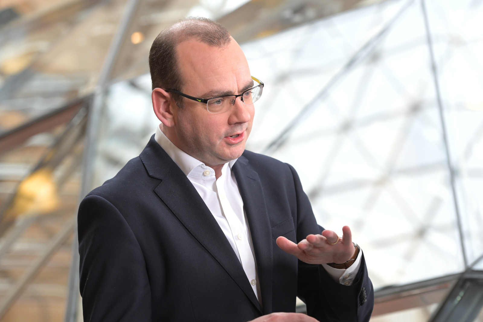 A professional man gestures while speaking, wearing a dark suit and white shirt, with a modern glass structure in the background.