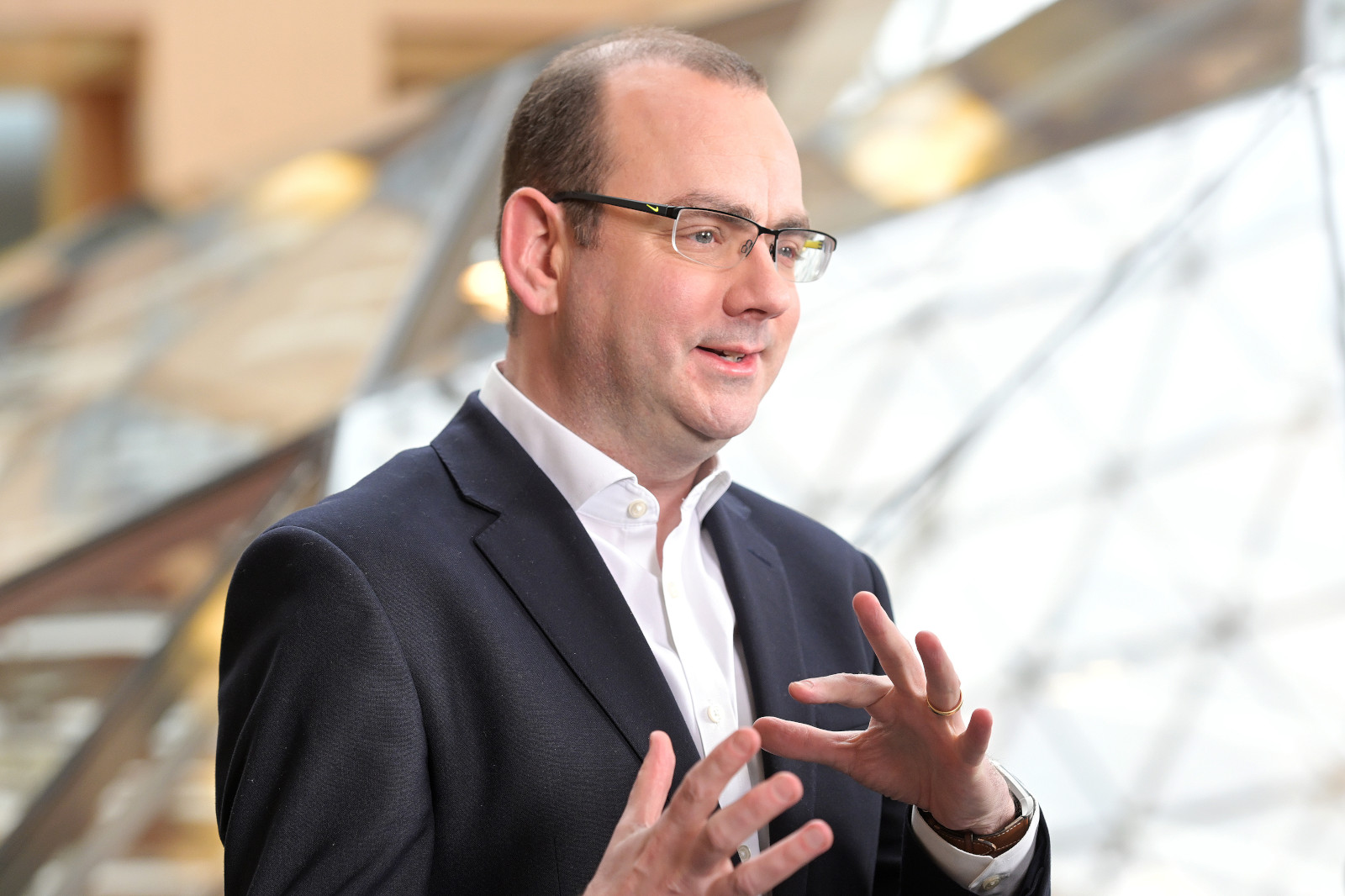 A person in a smart black suit gestures expressively while speaking, set against a modern, geometric background.