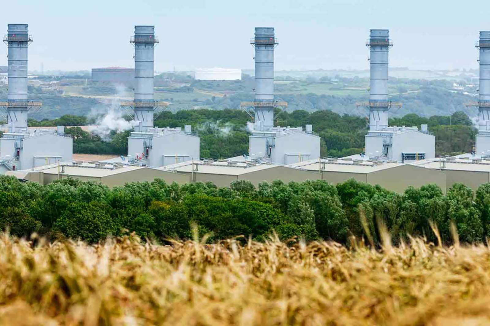 A view of industrial power plants with tall chimneys, surrounded by green trees and golden fields in the foreground.