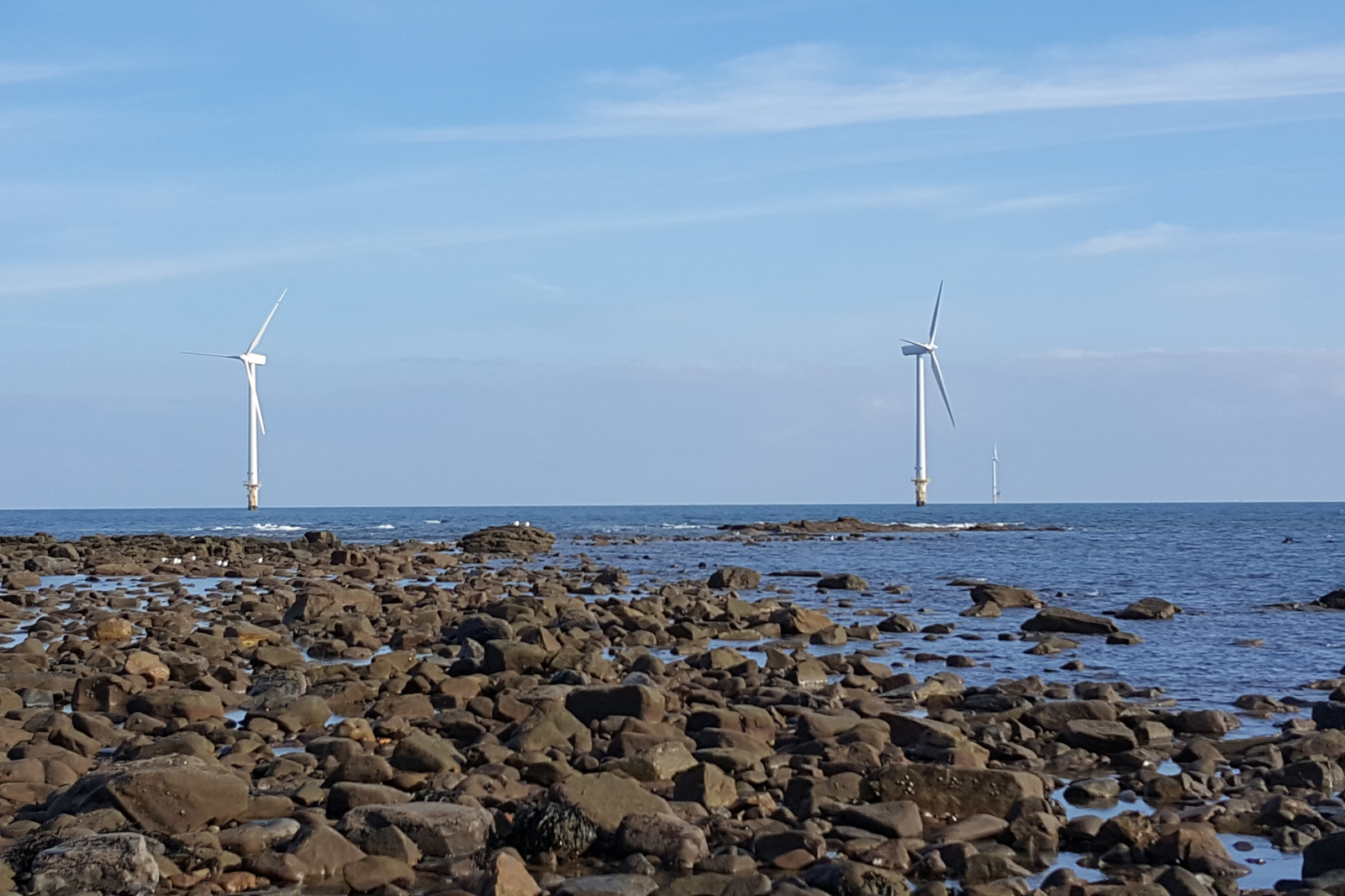 A scenic view of the sea with three wind turbines on the horizon, surrounded by rocky shoreline and blue sky.
