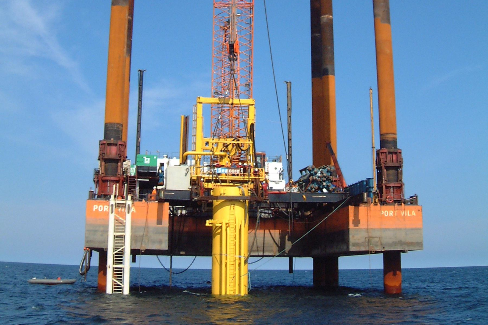 A large offshore oil rig with orange and yellow structures above the water, surrounded by calm blue sea and blue sky.
