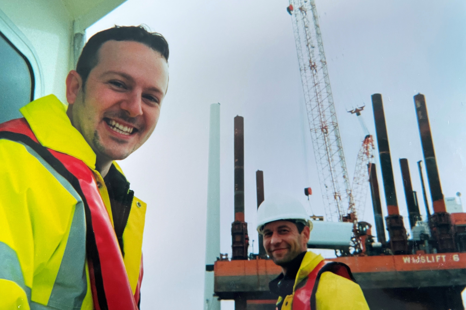 Two workers in safety jackets pose on a construction site with cranes and equipment in the background.