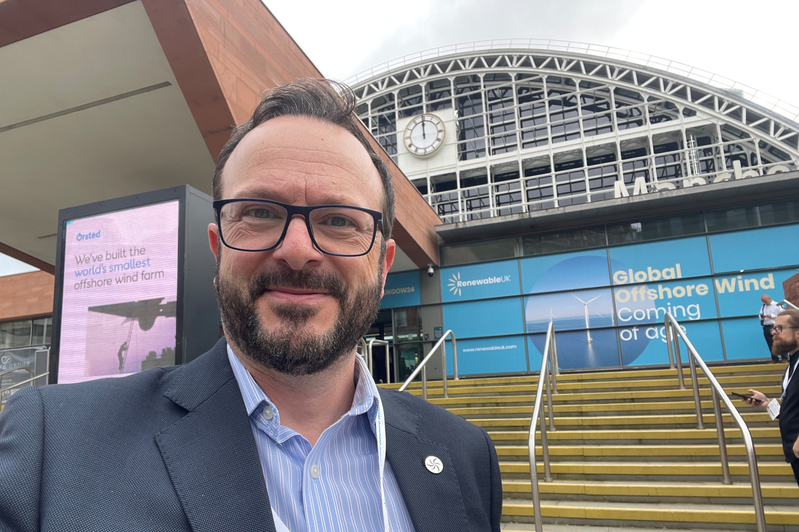 A person in a suit stands in front of a modern building featuring digital advertising for offshore wind farm initiatives.