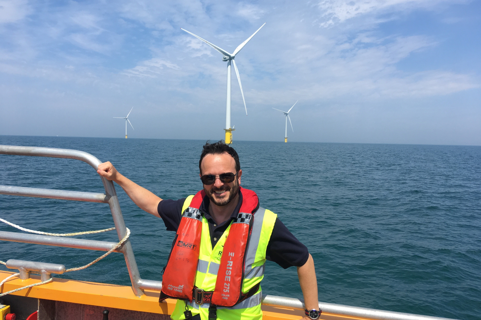 A person wearing a life jacket poses on a boat with offshore wind turbines in the background against a blue sky.