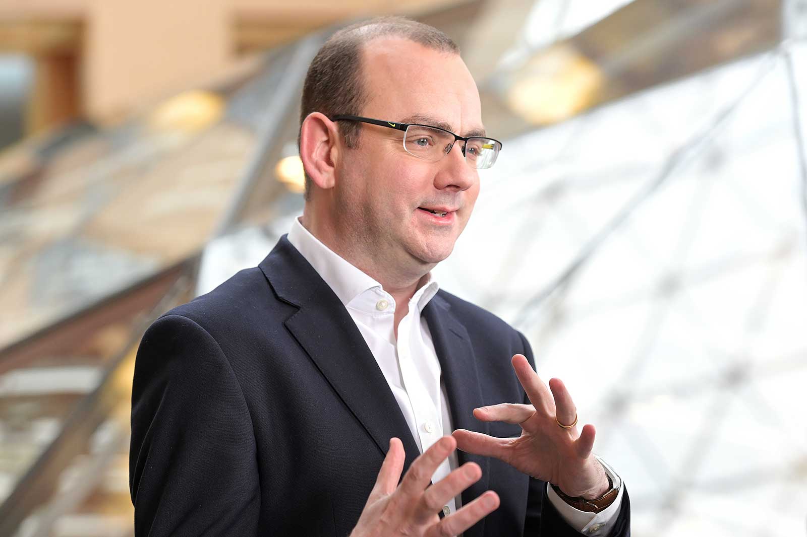 A person gestures while speaking, wearing a dark suit with a white shirt. A modern architectural backdrop is visible.