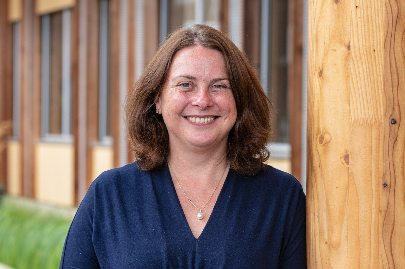 A woman with shoulder-length hair stands beside a wooden post in an outdoor setting, dressed in a navy blue top.