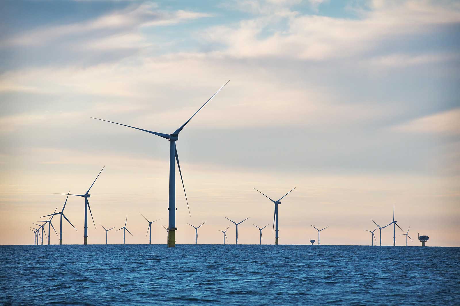 A row of offshore wind turbines stands tall against the horizon, with a calm sea and a pastel sky.