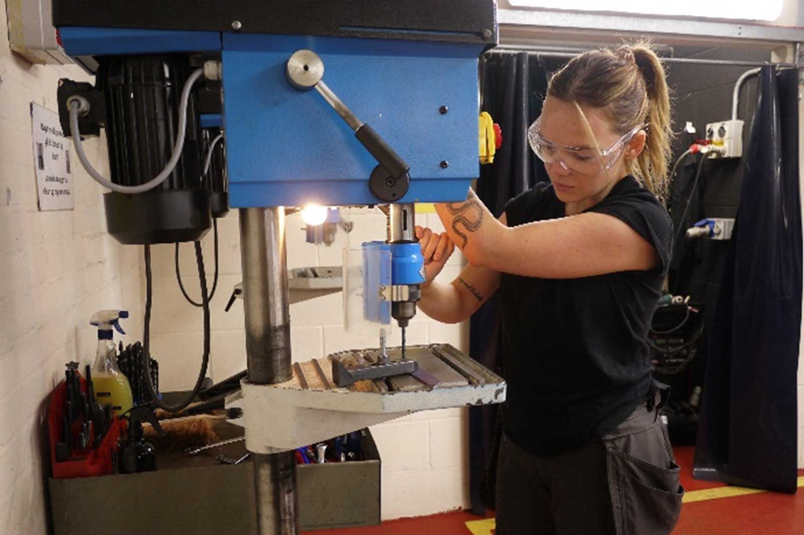 A person operates a blue drill press in a workshop, focusing on a metal piece on the machine's table.