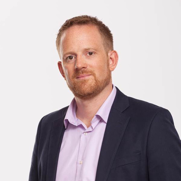 A man in a dark suit with a light purple shirt stands confidently against a plain background.