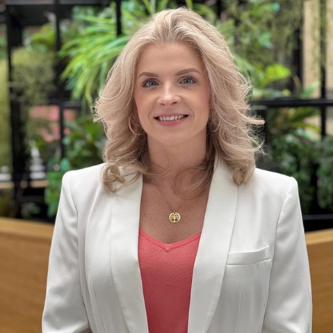 A woman with wavy blonde hair wearing a white blazer, standing against a lush green background. Portrait of Corinne Barry.