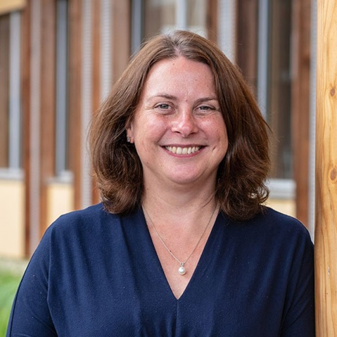 A woman with shoulder-length brown hair, wearing a navy blouse and a pearl necklace, stands near wooden architecture.