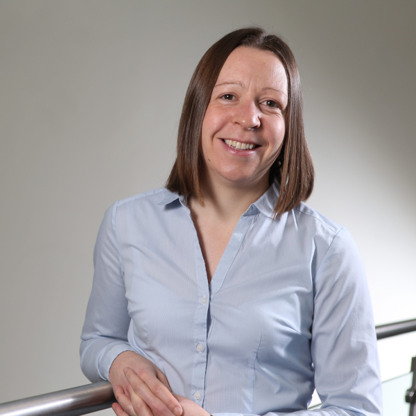 A woman in a blue button-up shirt stands by a railing, with a neutral background.