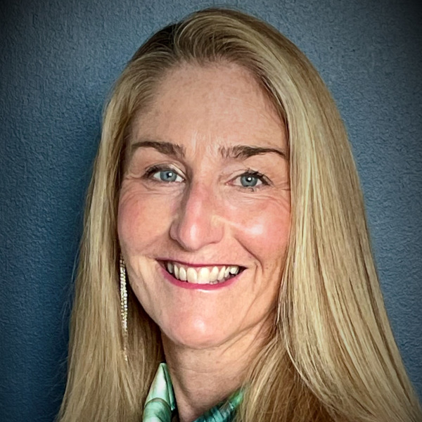Smiling woman with long blonde hair and blue eyes against a dark blue background, wearing a dangling sparkly earring and green scarf.