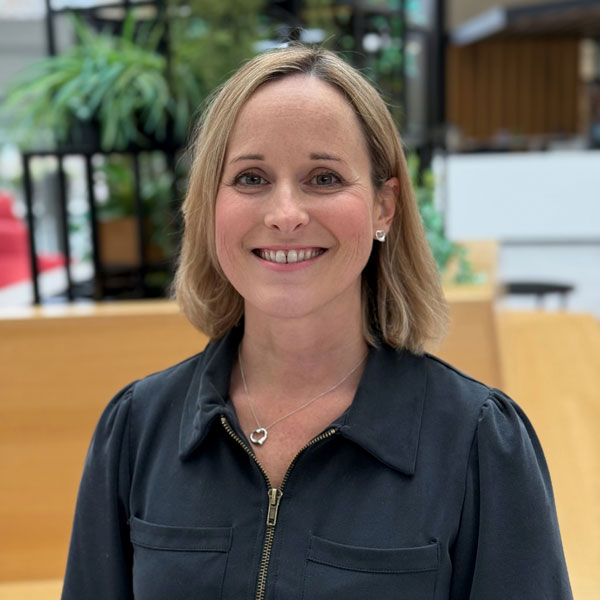 A woman with shoulder-length blonde hair is standing in a modern interior with plants and wooden elements in the background.