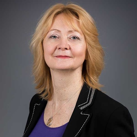 A woman with blonde hair, wearing a black jacket over a purple top, standing against a grey background.